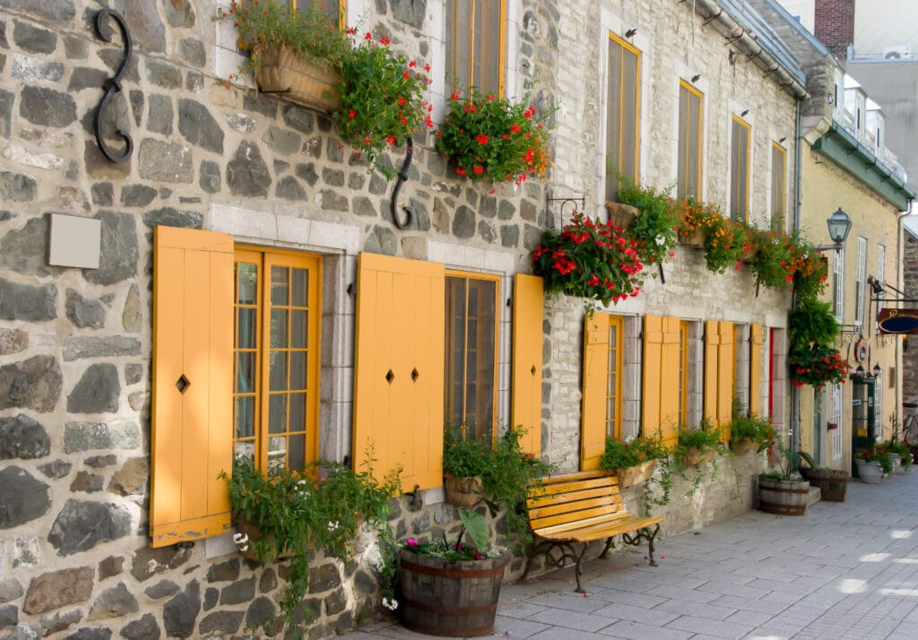 A street in Quebec City with stone houses. The houses have yellow shutters and colorful flowers at the windows. Quebec City is a port of call on Stitchers' Escapes 2023 fall needlework cruise.